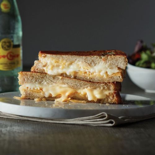 A gooey grilled cheese sandwich stacked on a plate, with melted cheese oozing out, next to a salad and a drink.