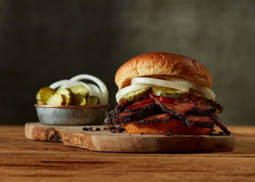 A close-up of a smoked brisket sandwich with pickles and onions, served on a wooden board with a side of pickles.
