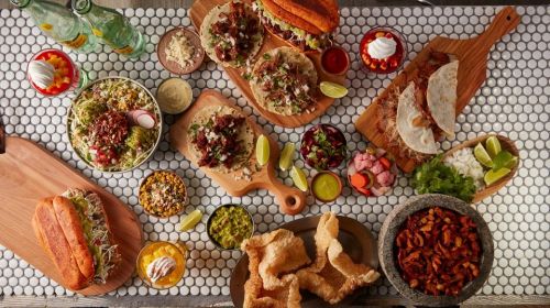A vibrant spread of tacos, salads, drinks, and snacks on a tiled table, showcasing a variety of Mexican dishes.