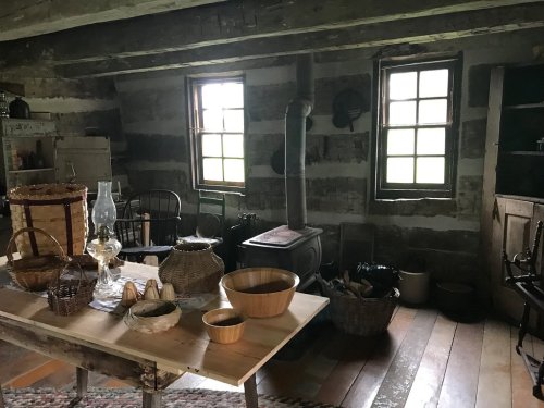A rustic interior of a cabin featuring wooden furniture, baskets, a stove, and natural light from two windows.