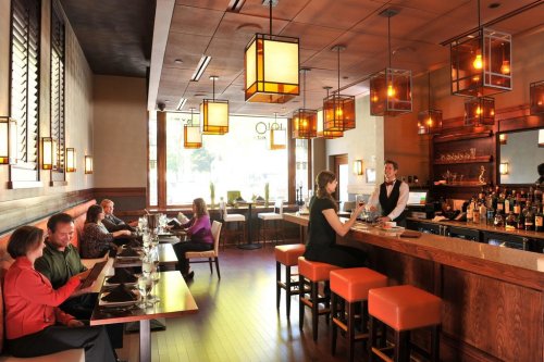 A modern restaurant interior with warm lighting, patrons dining, and a bartender serving at the bar.