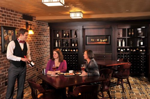 A waiter serves wine to two women at a cozy restaurant table, surrounded by a wine rack and warm lighting.
