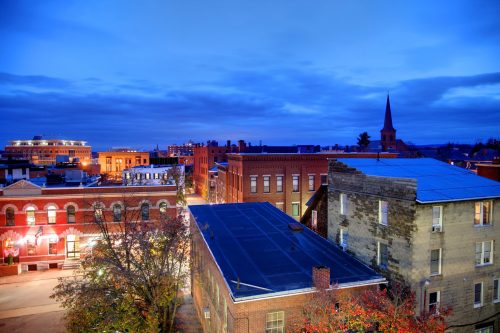 A twilight cityscape featuring historic buildings, a church steeple, and a cloudy blue sky.
