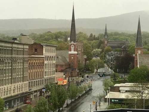A rainy view of a small town with church steeples, green trees, and historic buildings lining the street.