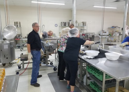 A busy kitchen with several people preparing food, wearing gloves and hairnets, and using various kitchen equipment.