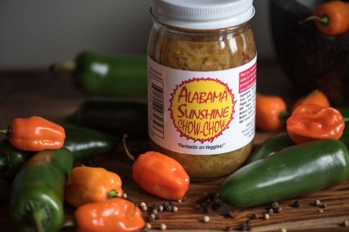 A jar of Alabama Sunshine Chow Chow surrounded by fresh peppers and spices on a wooden surface.