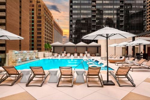 Rooftop pool area with lounge chairs and umbrellas, surrounded by tall buildings and a sunset sky.