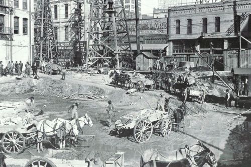 Historic construction site with horse-drawn carts, workers, and buildings in the background. Dust and activity abound.