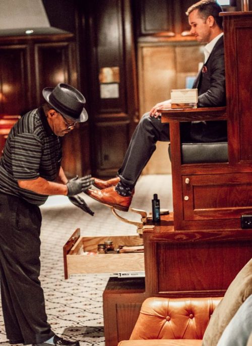 A man in a suit sits while another man polishes his shoes in a stylish, upscale setting.