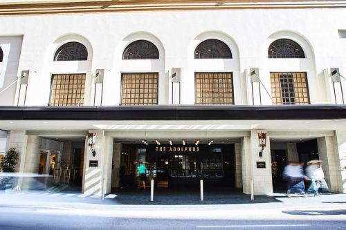 The entrance of The Adolphus hotel, featuring large windows and a stylish facade, with blurred figures walking by.