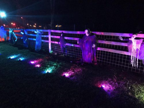 A dark scene featuring ghostly figures hanging on a fence, illuminated by colorful lights on the ground.