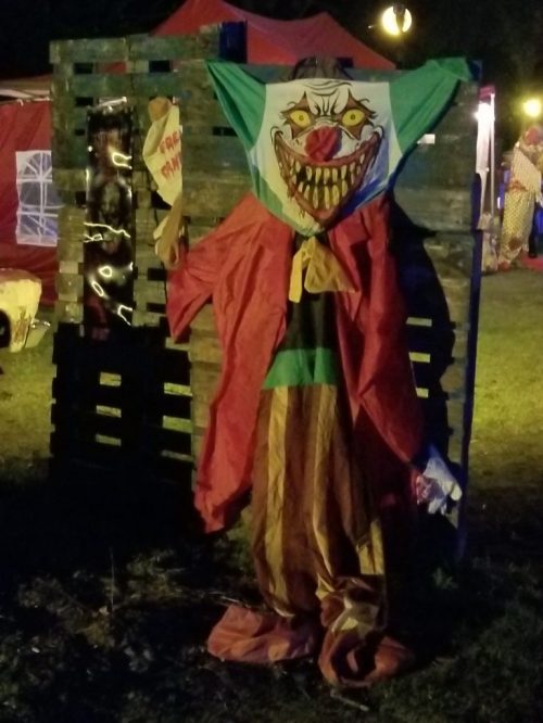 A creepy clown figure hangs on a wooden backdrop, illuminated by dim lights in a dark outdoor setting.