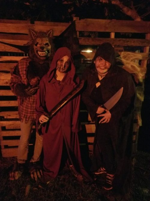 Three figures in Halloween costumes: a wolf mask, a hooded figure with a skull face, and a cloaked figure with a knife.