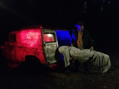 A figure in a dark outfit holds a wrapped body near an old truck illuminated by red and blue lights in a dark setting.