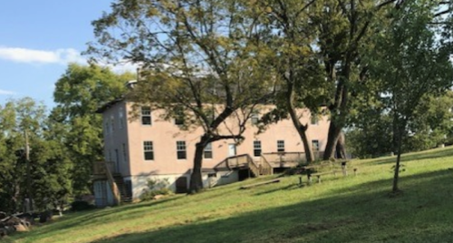 A two-story house on a grassy hill, surrounded by trees under a clear blue sky.