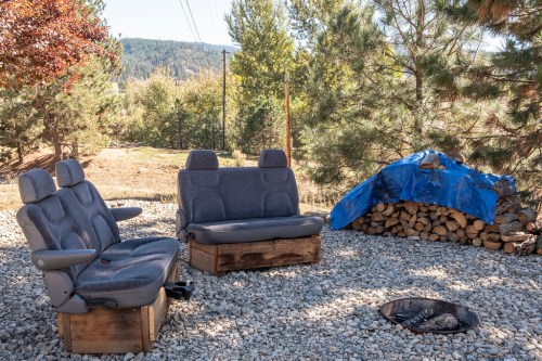 Two cozy, gray upholstered seats sit on gravel near a fire pit and a stack of firewood covered with a blue tarp.