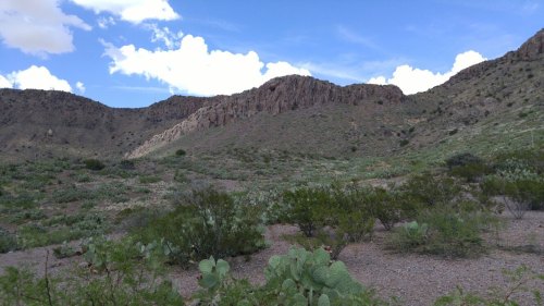 A desert landscape featuring rolling hills, sparse vegetation, and a blue sky with scattered clouds.