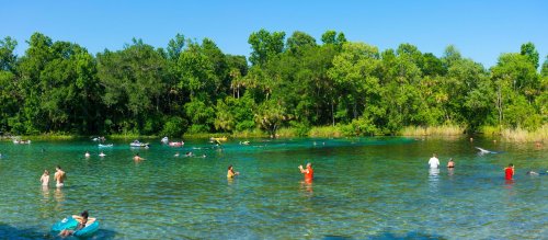A sunny day at a clear blue lake with people swimming and enjoying the water surrounded by lush green trees.