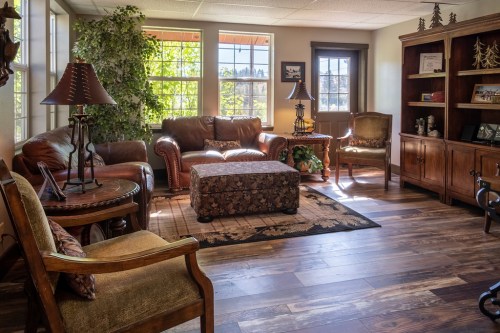 Cozy living room with leather sofas, a coffee table, and a wooden bookshelf, featuring large windows and warm lighting.
