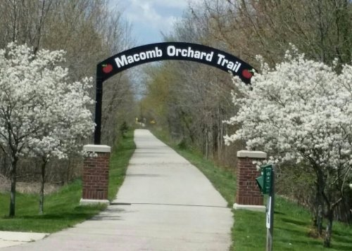 Archway sign for Macomb Orchard Trail, flanked by blooming trees, leading down a paved path through a natural setting.