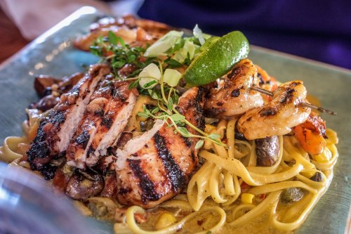 Grilled chicken and shrimp served over fettuccine with vegetables and a lime wedge on a blue plate.