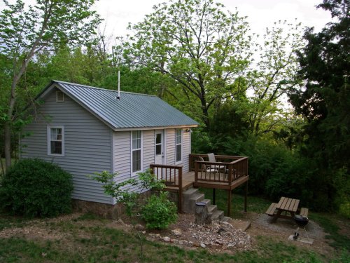 A small cabin with a metal roof, surrounded by trees, featuring a deck and outdoor seating area.