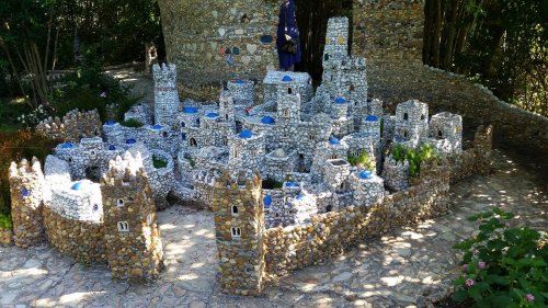 A miniature castle made of stones and pebbles, featuring towers and blue accents, set in a lush garden.