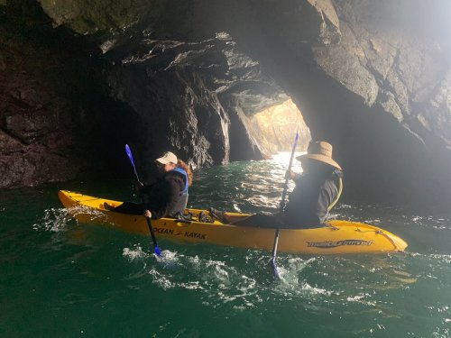 Two people in a yellow kayak paddling through a rocky cave with water reflecting light.