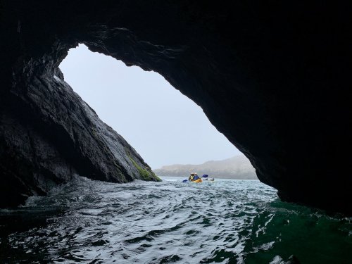 A kayaker paddles through a rocky cave, with waves and misty scenery visible beyond the archway.