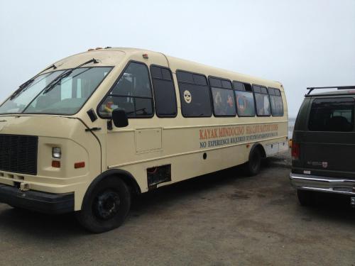 A yellow bus with "Kayak Kyndo" signage parked near a body of water, with a gray van nearby.