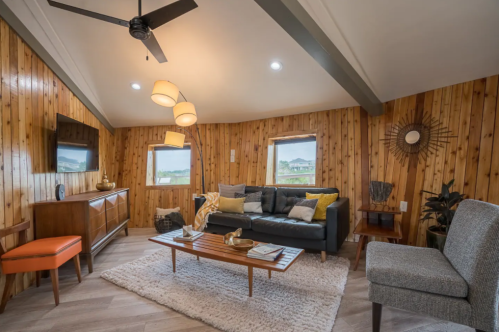 Cozy living room with wooden walls, modern furniture, a ceiling fan, and large windows showcasing a scenic view.