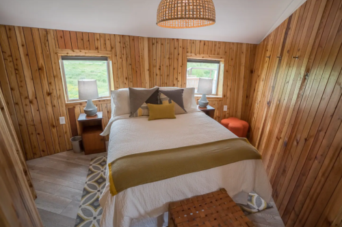 Cozy bedroom with wooden walls, a double bed, two lamps, and a small orange stool by the window.
