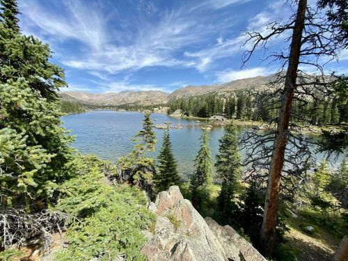 A serene lake surrounded by lush trees and mountains under a blue sky with wispy clouds.