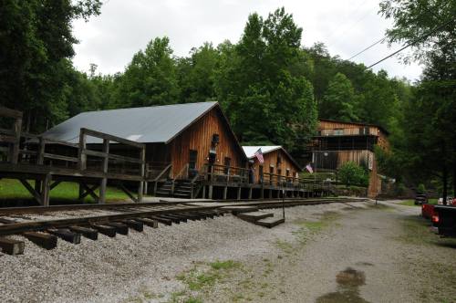 A rustic wooden building with a metal roof, surrounded by trees and a gravel path, with railroad tracks in the foreground.