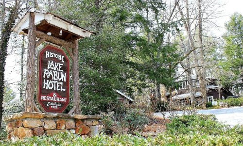 Sign for the Historic Lake Rabun Hotel & Restaurant, surrounded by trees and greenery, with a rustic wooden structure in the background.