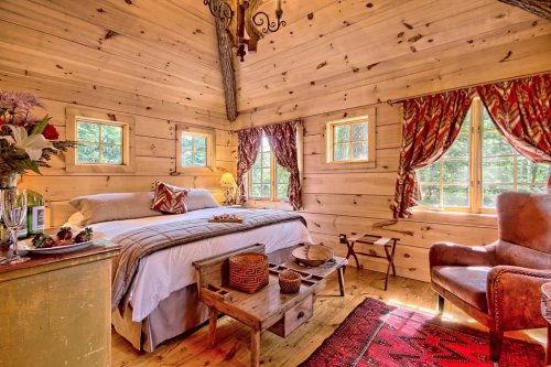 Cozy wooden bedroom with a bed, decorative curtains, a chair, and a small table adorned with flowers and snacks.
