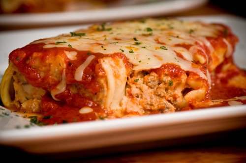 A close-up of a cheesy lasagna slice topped with marinara sauce and garnished with parsley on a white plate.