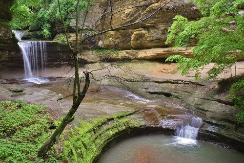 A serene landscape featuring two waterfalls cascading into a rocky pool, surrounded by lush greenery and cliffs.