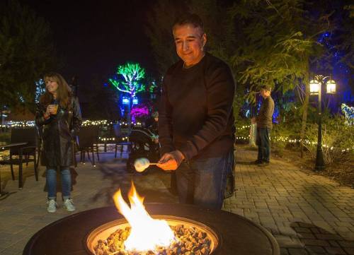 A man roasting marshmallows over a fire pit at night, with colorful lights and people in the background.
