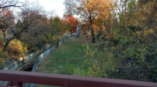 A view of a grassy area bordered by trees with autumn foliage, seen from a bridge.