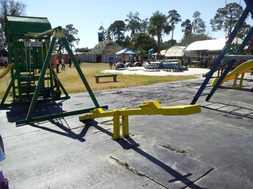 A playground with swings and a seesaw, set in a grassy area with people and tents in the background.