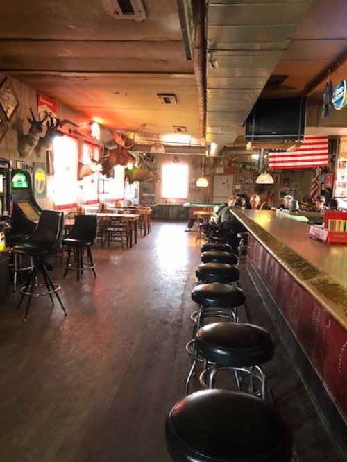 A rustic bar interior with empty stools, wooden tables, and decor including mounted animal heads and vintage signs.