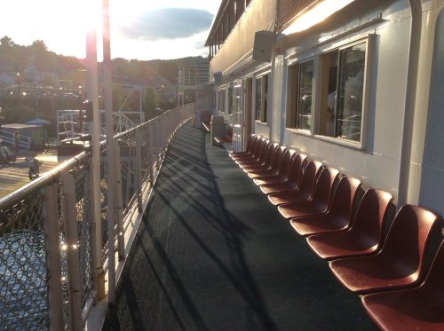 A sunlit deck of a boat with red chairs lined up along the railing, casting long shadows on the surface.