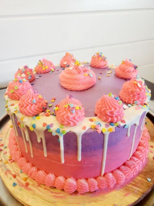 A colorful layered cake with pink and purple frosting, topped with swirls and colorful sprinkles.