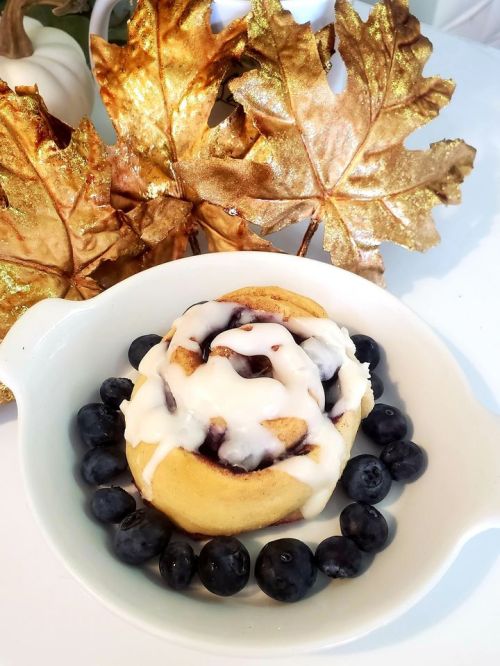 A cinnamon roll topped with icing, surrounded by fresh blueberries, with golden autumn leaves in the background.
