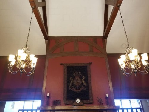 Interior view featuring a high ceiling with wooden beams, chandeliers, and a decorative wall hanging on a red background.