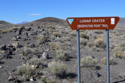 Sign for Lunar Crater Observation Point Trail, with rocky terrain and sparse vegetation under a clear blue sky.