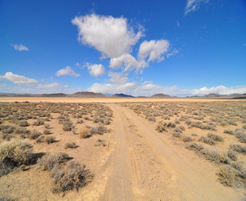 A dirt path leads through a sparse desert landscape under a blue sky with fluffy clouds and distant mountains.