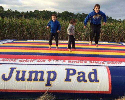 Three children joyfully jumping on a colorful inflatable jump pad in a field.