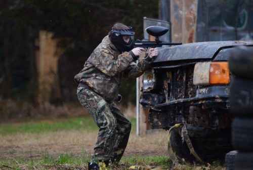 A person in camouflage gear aims a paintball gun while crouching behind an old, rusty vehicle in an outdoor setting.